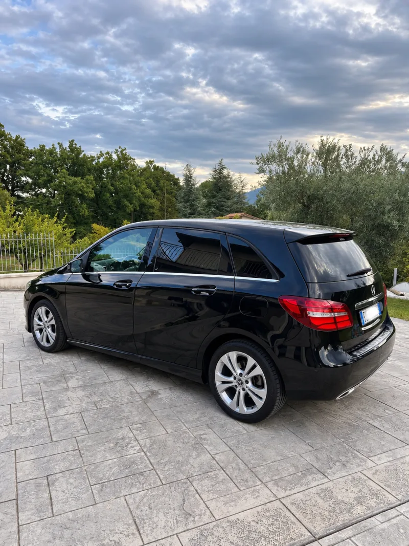 Die schwarze Mercedes B‑Klasse von hinten in Dreiviertelansicht, in einem Hof mit Olivenbäumen und Bergen