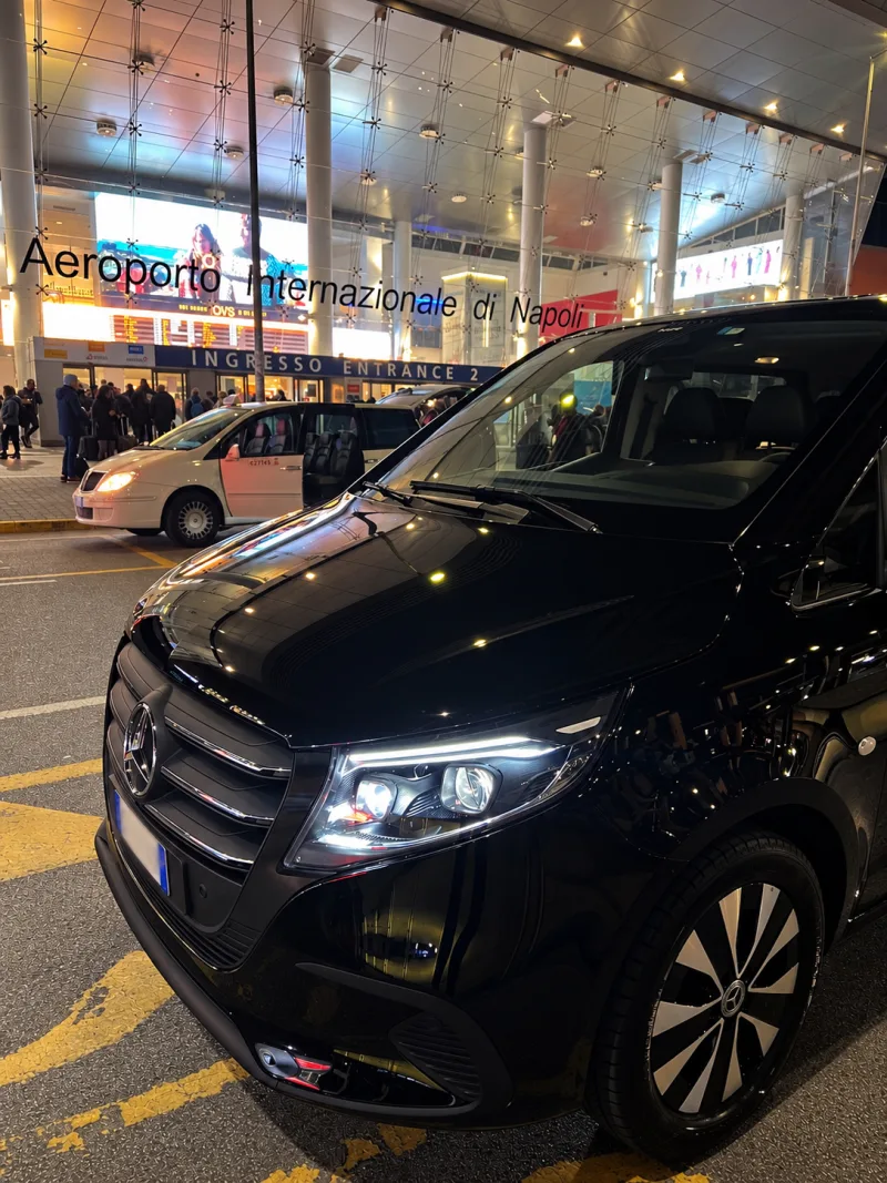 Der schwarze Mercedes Vito Extralong 2026 mit eingeschalteten Scheinwerfern vor dem Internationalen Flughafen Neapel, am Abend